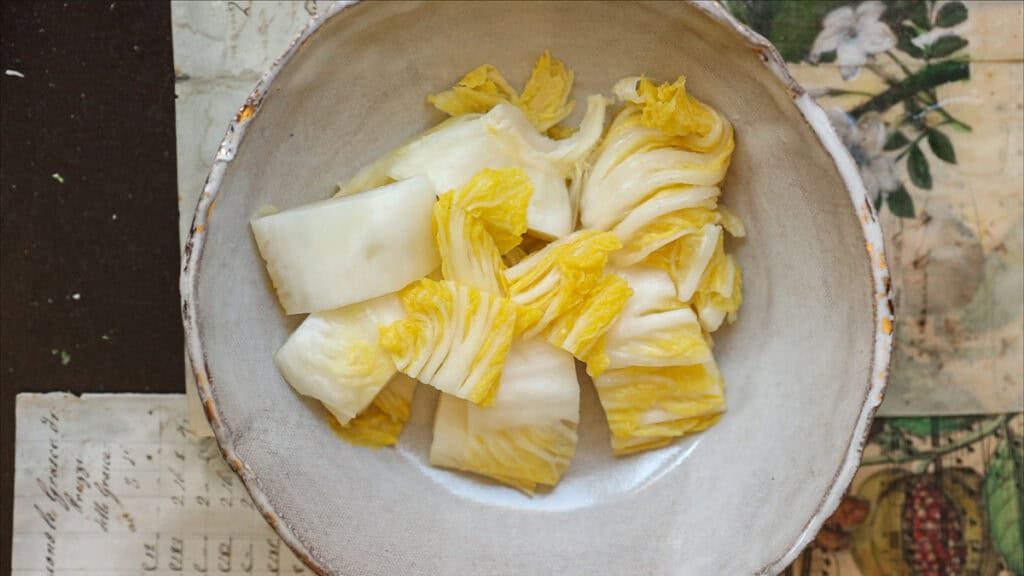 A bowl of Baek-Kimchi, or 백김치, rests on a table adorned with vintage floral and script-patterned papers. The napa cabbage is pale yellow and white, while the bowl exudes a rustic charm, perfectly capturing the simplicity of an easy white kimchi recipe.